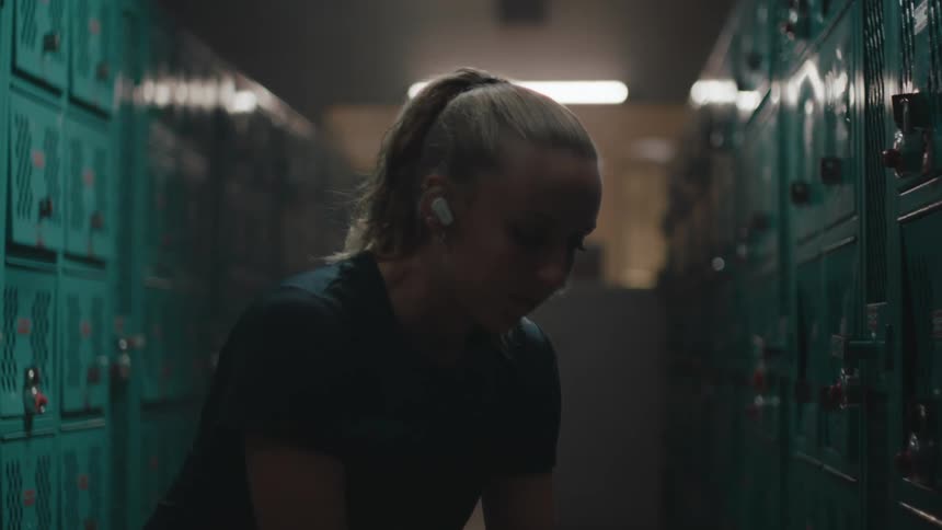 A young blonde woman is sitting in a place with many cabinets around her and is wearing white headphones