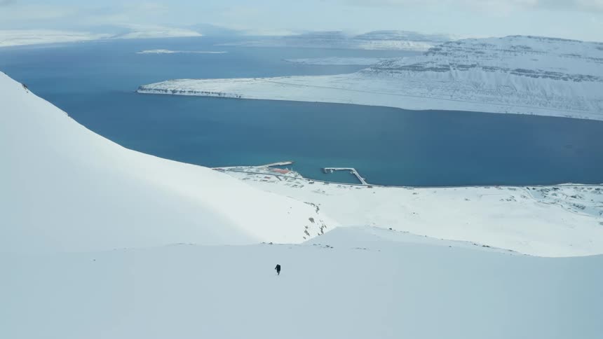 A person walking on a white mountain covered with snow and behind is a sea