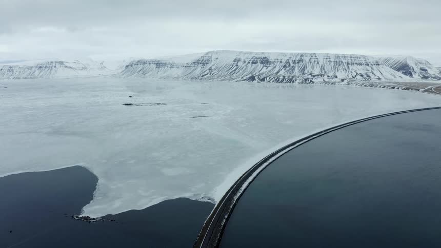 Aerial panorama of a long road crossing an icy sea and snowy mountains