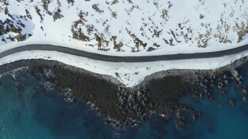 Aerial panorama of a winding road surrounded by snowy mountains and next to a blue sea