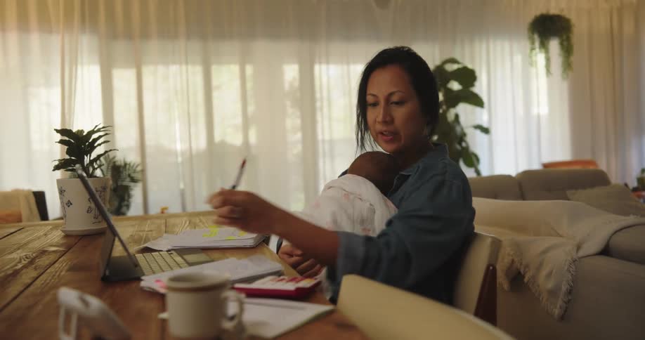 Mother holds baby during video call while working at table in home