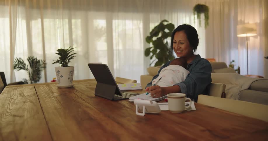 A mother sitting at her dining room table holding her infant baby in her arms in front of tablet