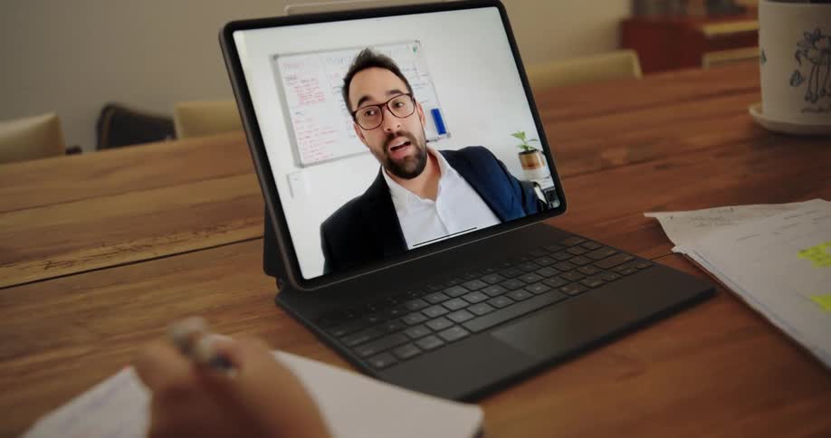 A man is seen talking on a laptop computer screen as the person sitting at the table is writing in a notebook