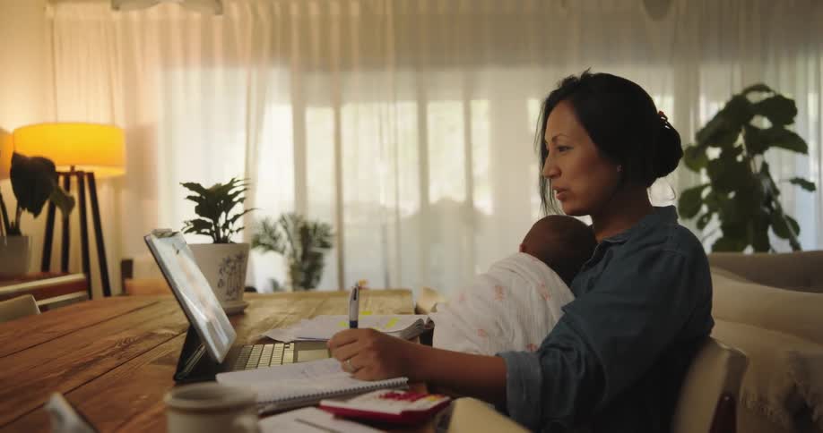 A woman is sitting at a table in talking to a man on a laptop computer as she holds her baby and writes in a notebook