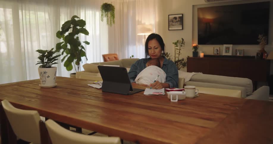 Awoman sits at a large wooden table in a living room in front of a laptop she is holding a baby as she writes on a pad of paper in front of her and smiles and responds to someone on the laptop