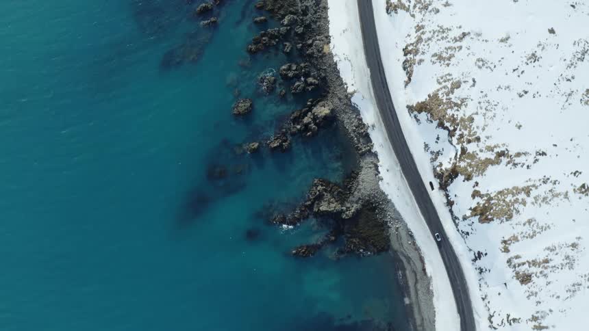 A car driving on a road between a snowy mountain and the ocean