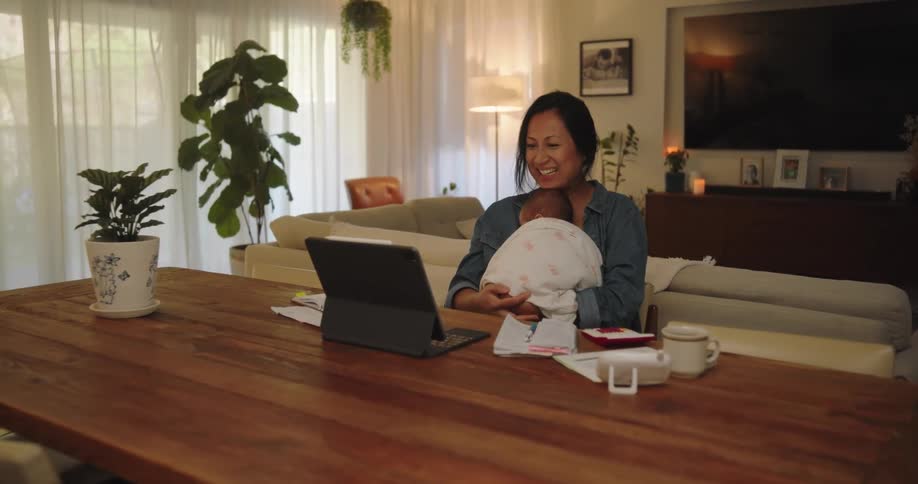 Young mother and newborn baby sitting at a long wooden table in front of a tablet while the mother smiles and talks to someone on the screen