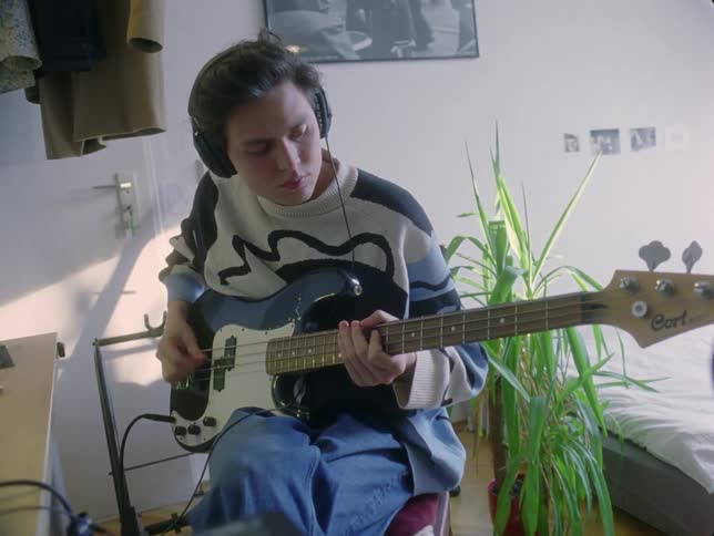 A young person wearing headphones plays a guitar while sitting bobbing his head composing music at his computer