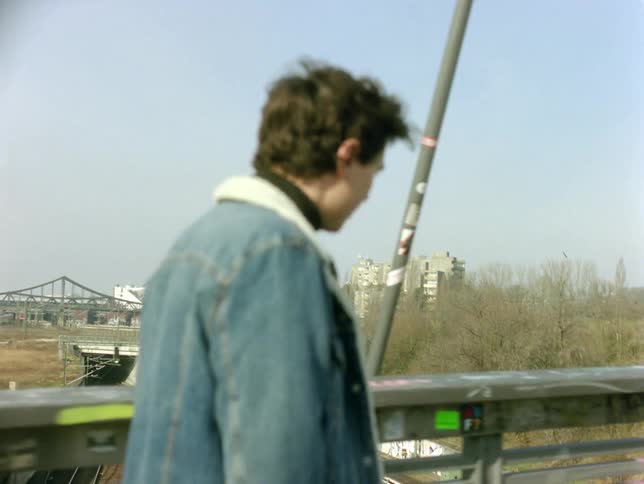 Young man walks across bridge over highway and train tracks with buildings in the background