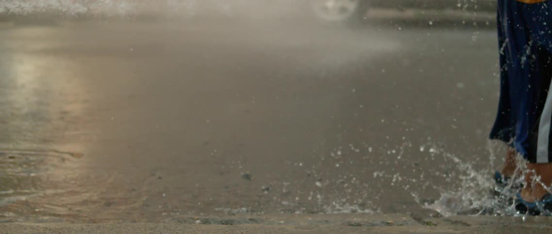 Legs of a child jumping into a puddle of water on the street and next to a burst fire hydrant releasing water