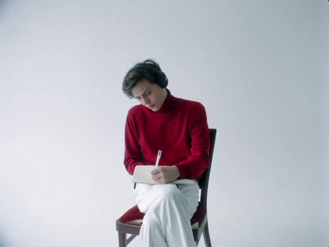 Young man sitting on a chair with his legs crossed writing in a notebook and using headphones