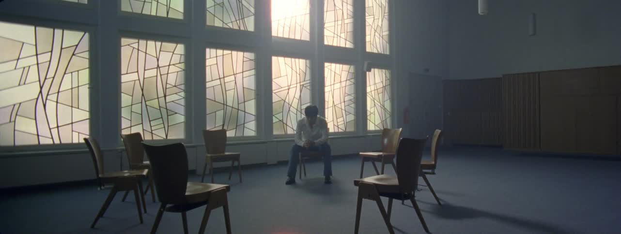 A young asian man sits alone in a room with several empty chairs