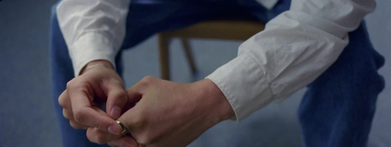 A young man clasping their hands together wearing a white shirt and blue pants with a ring on one finger
