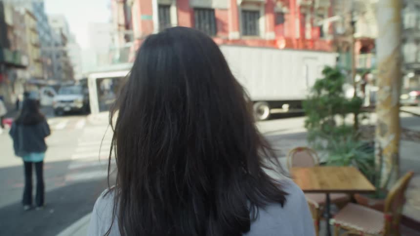 Perspective of a blackhaired woman seen from behind walking along a sidewalk in an urban city in daylight