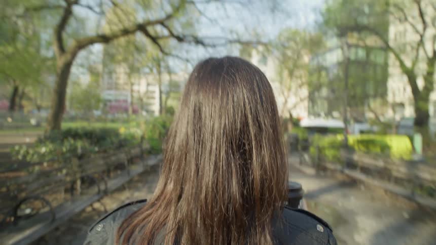 Point of view of a blackhaired woman holding a disposable coffee cup walking on the sidewalks of a park with trees around her