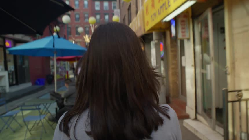 Point of view of a blackhaired woman walking from behind down a narrow street with commercial stores in a metropolis