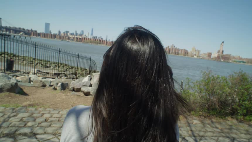 A woman brushes back her hair while sitting at the waterfront looking across to New York City