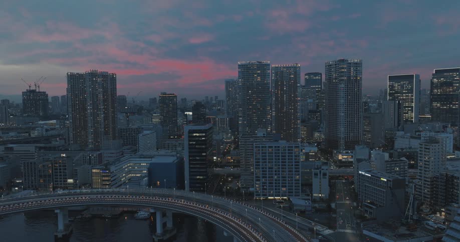 Aerial view of Tokyo's Rainbow bridge loop during magic hour revealing a breathtaking nighttime panorama of the city adorned with multicolored skyscrapers against a deep blue sky dotted with few clouds