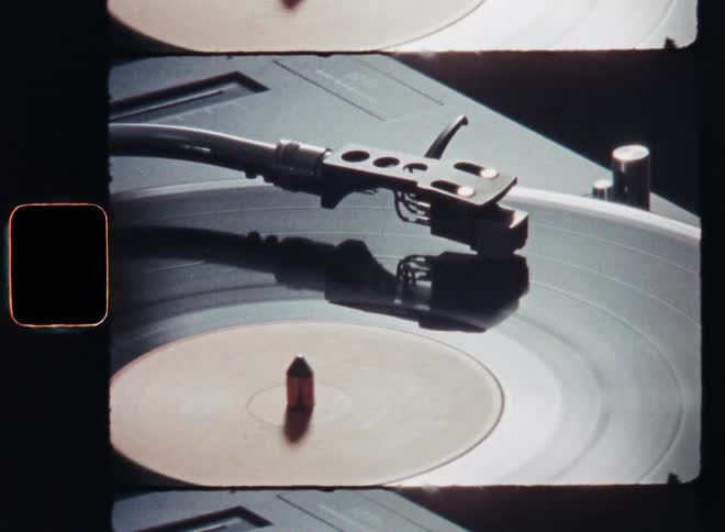 Old super 8mm film footage of a vintage record player or turntable with a sound needle on a vinyl EP or album spinning or turning in motion with light reflection as the sun reflects on the glossy surface of the analog media