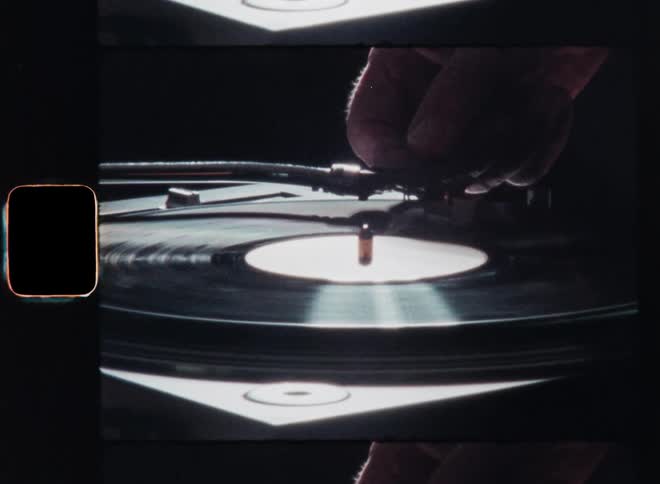The hand of a person or DJ artist sets the arm of a record player carefully down on a spinning vinyl record as he plays the next track on the stage of a live show event as the lights reflect on the shining surface in front of a dark background