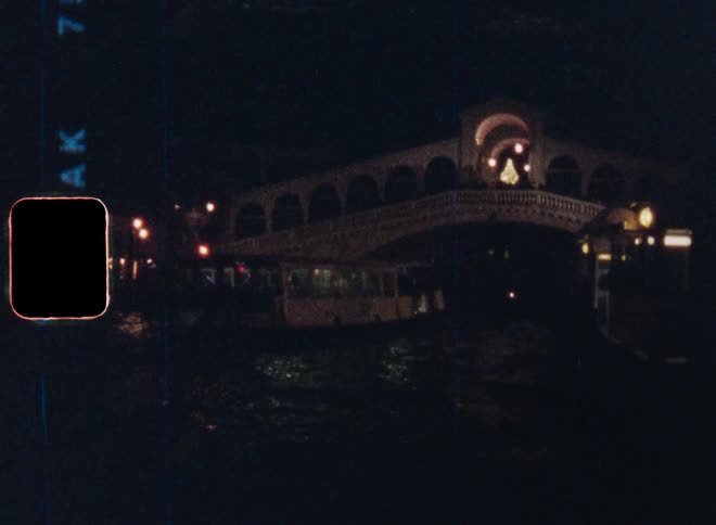 A nighttime scene on Super 8mm film of a ferry boat driving on a river or canal underneath the famous Rialto Bridge illuminated by lights in Venice Italy
