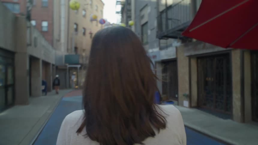 Perspective of a woman with black hair seen from behind walking through an alley with businesses in an urban city