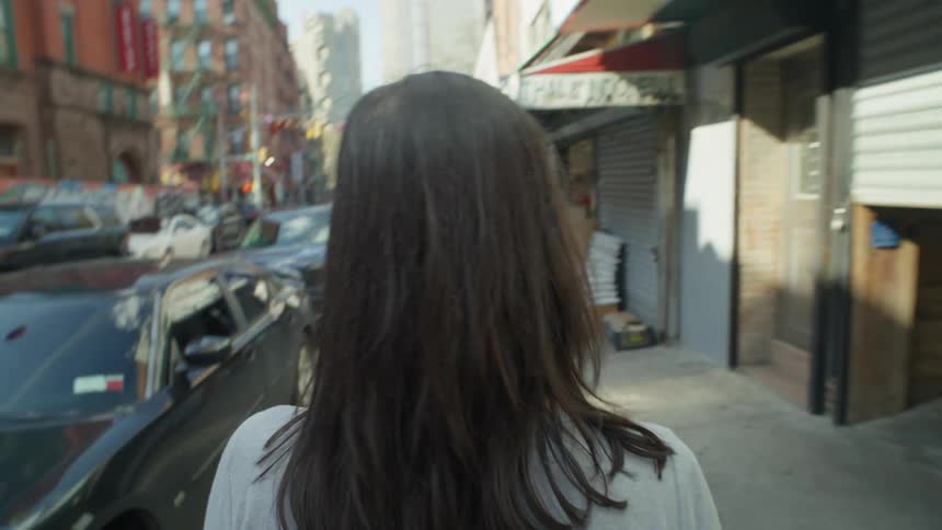 Perspective of a woman with black hair seen from behind walking on the sidewalk of a city with parked cars and commercial stores