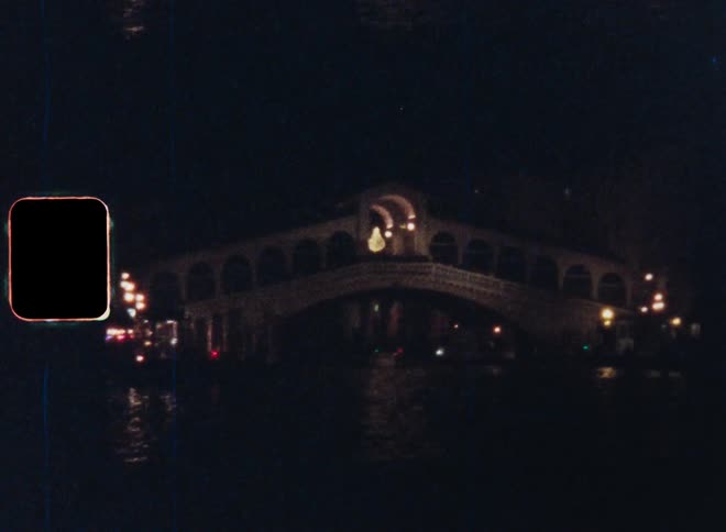 Point of view of a boat on a lake or river passing through a canal approaching the illuminated Rialto bridge in Venice city in the dark at night on Super-8 film