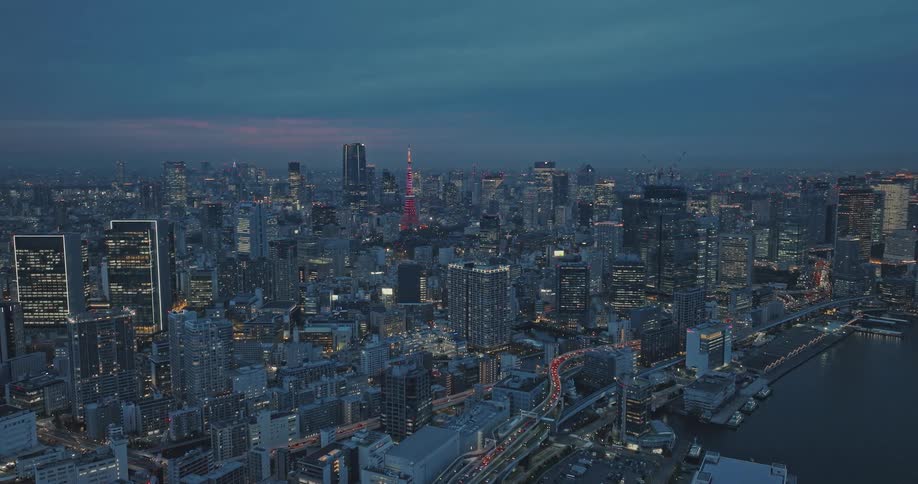 Aerial nighttime panorama of the Tokyo Tower adorned with multicolored skyscrapers against a deep blue sky dotted with clouds