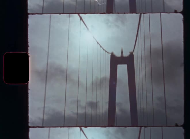 Vehicle crosses a monumental hanging or suspension bridge as an impressive sight during a sightseeing road trip with a vintage car as the Super 8mm camera zooms in onto the bridge tower architecture while driving on a cloudy day with sun