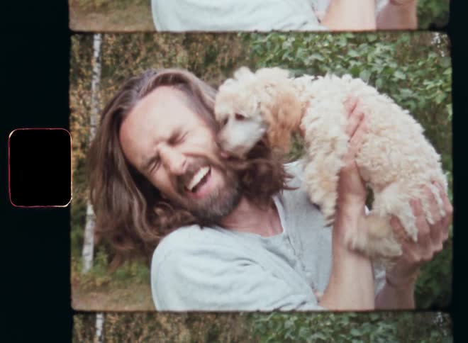 Young brown-haired man holding his cute poodle puppy in his arms as the small dog licks his face and he laughs while the animal expresses his love to his caring and loving pet owner on retro Super 8 film footage