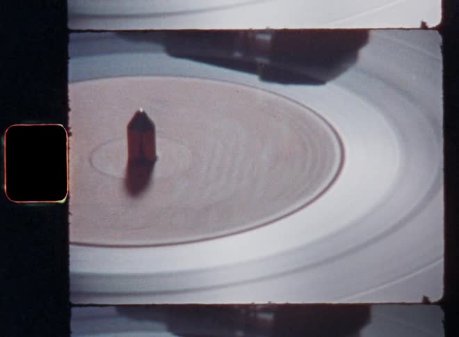 A vinyl record spins on a turntable as the smooth and reflective surface is reflecting the studio lights on the stage of a night club and the dance music is played loud on Super 8mm film footage with upward tilting camera movement