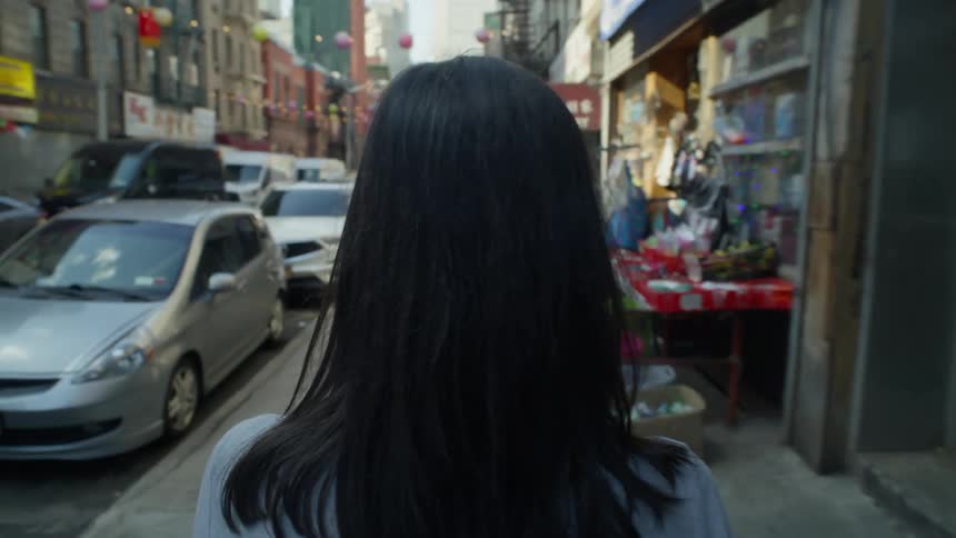 A woman with dark hair wearing a white shirt walks along the sidewalk passing parked cars and buildings in the city