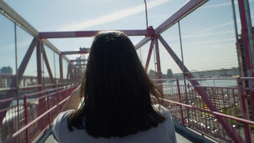 Perspective of a woman with black hair seen from behind walking on a bridge in a metropolis