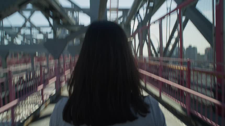 Back of the head of a woman with long hair walking on a bridge with a metal structure and a metropolis in the background