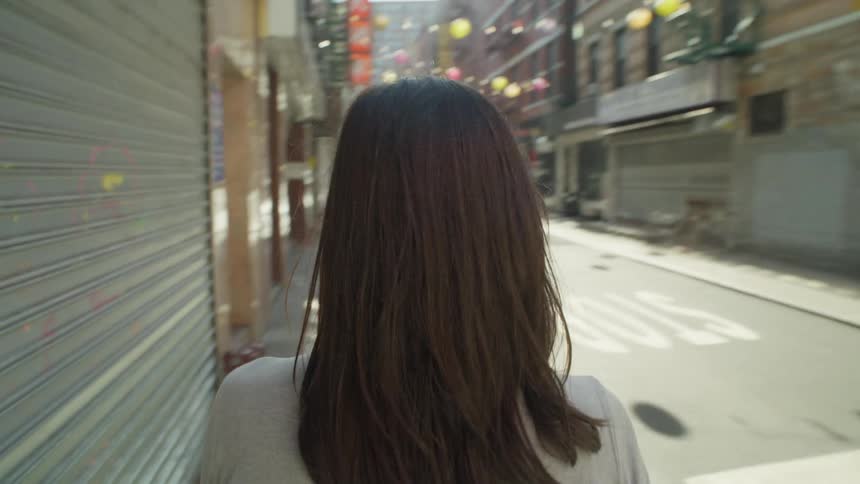 Perspective of a woman with black hair seen from behind crossing the street and walking along the sidewalk of a city