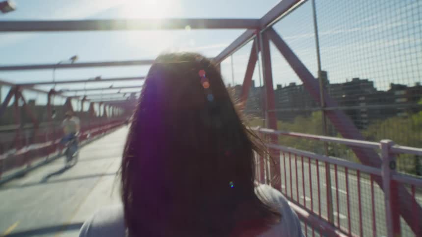 Perspective of a blackhaired woman seen from behind walking on an overpass in an urban city
