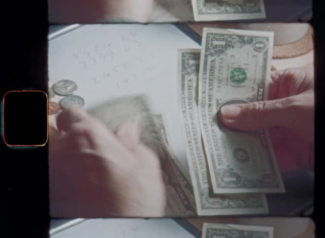 Hands of a man counting several dollar bills on a table with coins and then making a note of what he made or earned during the bookkeeping or cash check of his small family business store with coffee mug and calculator on the wooden table