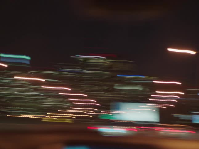 A nighttime cityscape with illuminated buildings and cars on a road seen from a moving vehicle