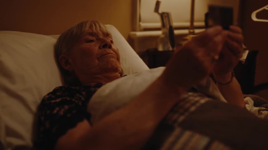 As the elderly woman lies in bed she gazes at a photo in her hands her emotions stirring within her