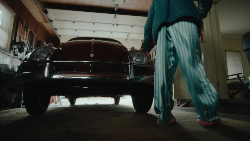 An elderly woman walks around a car parked in the garage