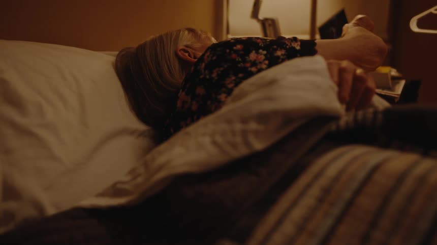 An elderly woman is lying on a bed and looking at a photo and then turns to the side to sleep
