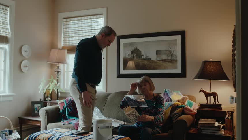 A man helps his wife to adjust a magnifying glass for reading in the living room