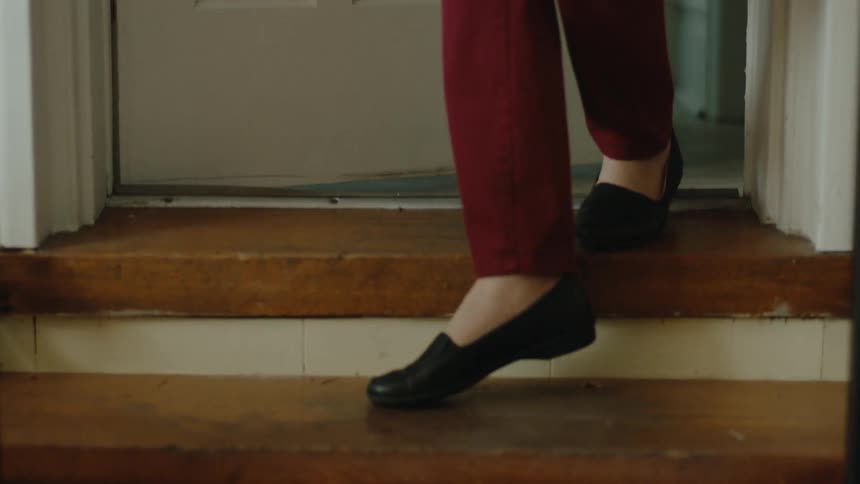 A woman in red pants and black shoes is stepping down a wooden staircase indoors