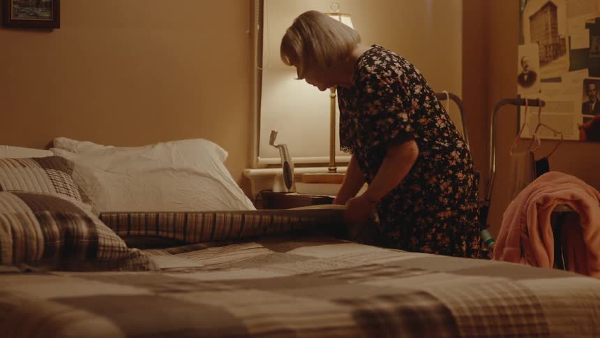 An elderly woman turns down her bed sheets and sits on the bed