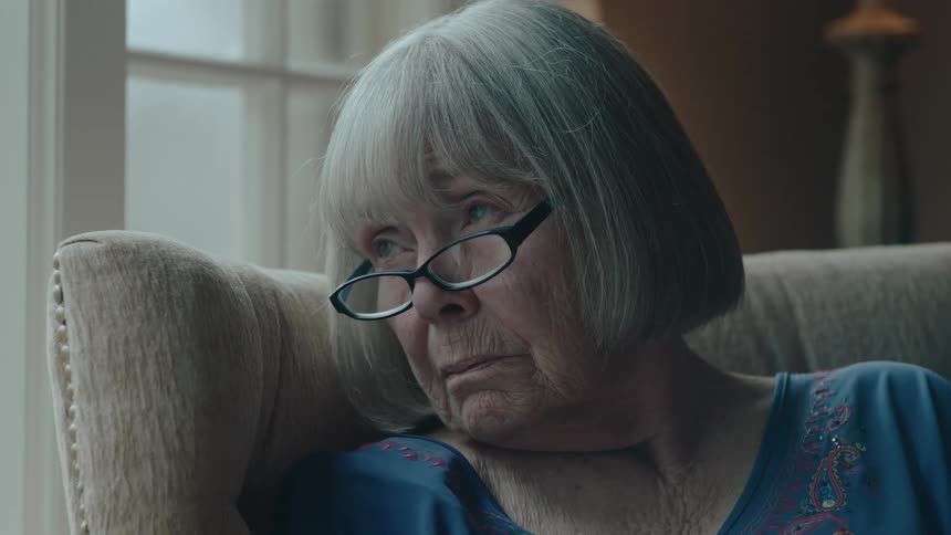 The face of an old lady wearing reading glasses and she is sitting in an armchair