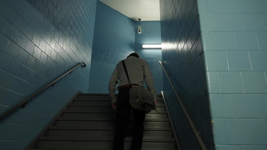 A man with long hair wearing a shirt black pants and a shoulder bag ascends a staircase near the blue tiled walls