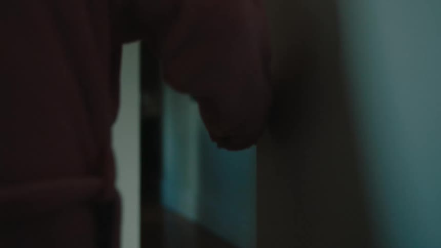 Closeup of the back of an elderly woman in pink pajamas walking down the hallway of the house leaning against the wall