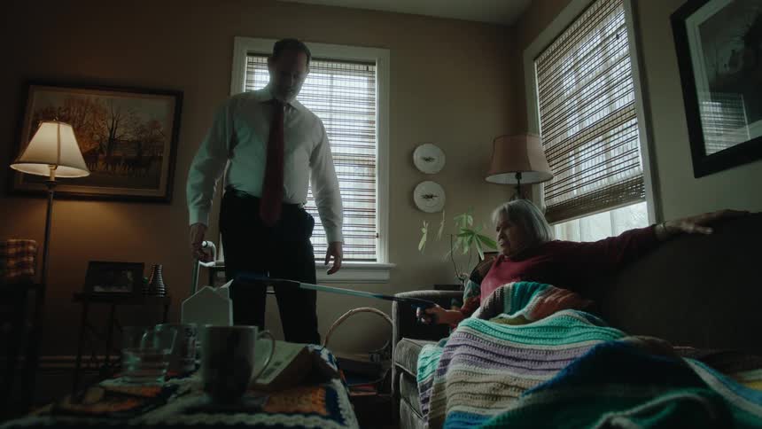 Man standing trying to pick up an object on the table and a lady lying on the sofa next to him fighting with him while holding a metal tongs