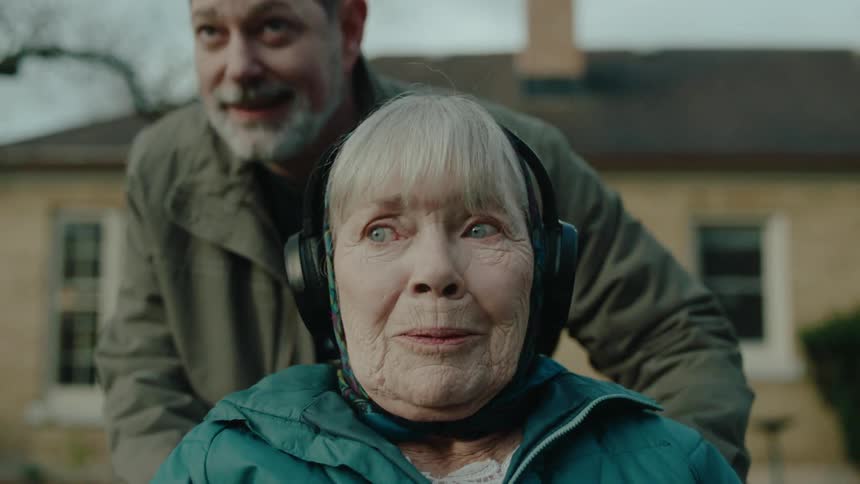 Man carrying an elderly woman in wheelchair with headphones who is emotional